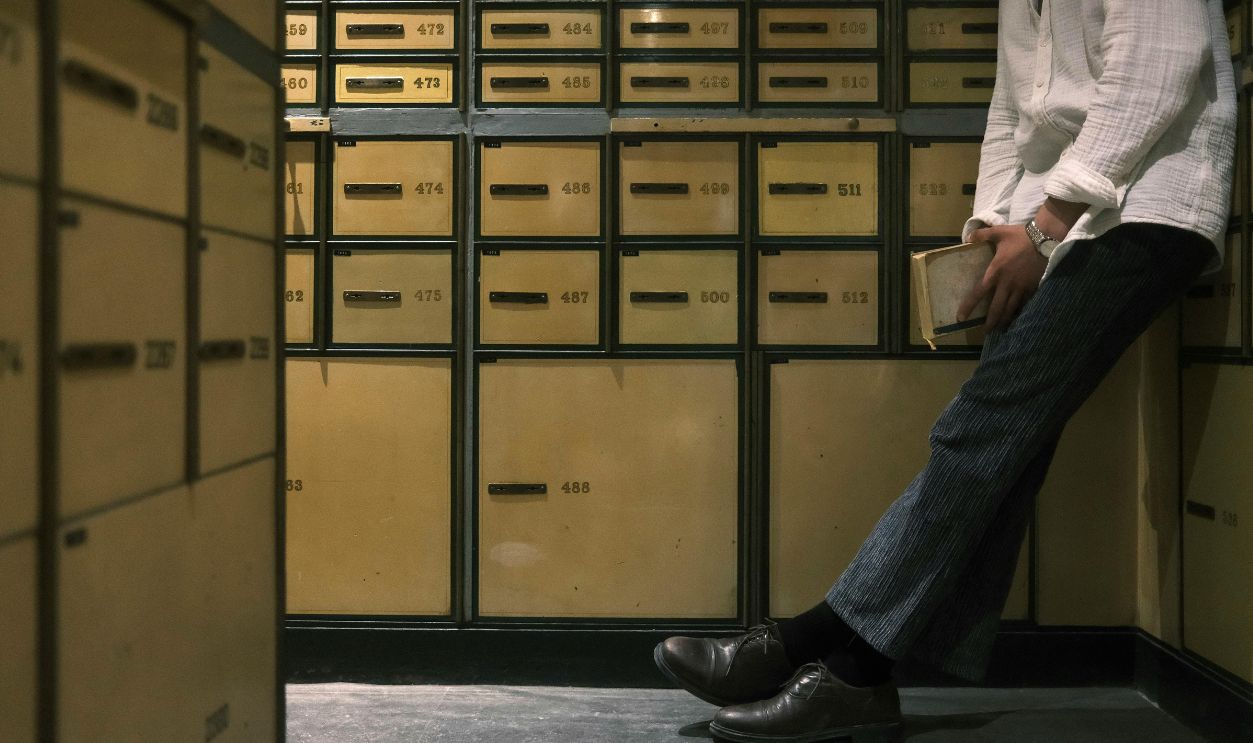 Man standing and leaning against mailboxes, holding a book in a dim-lit interior