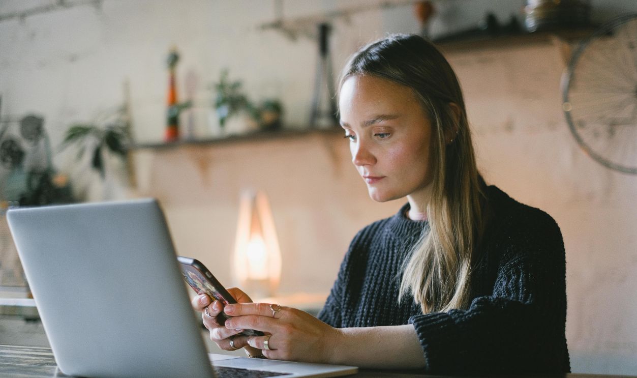 Young woman focusing on her smartphone and laptop in a cozy indoor workspace. Ideal for illustrating remote work