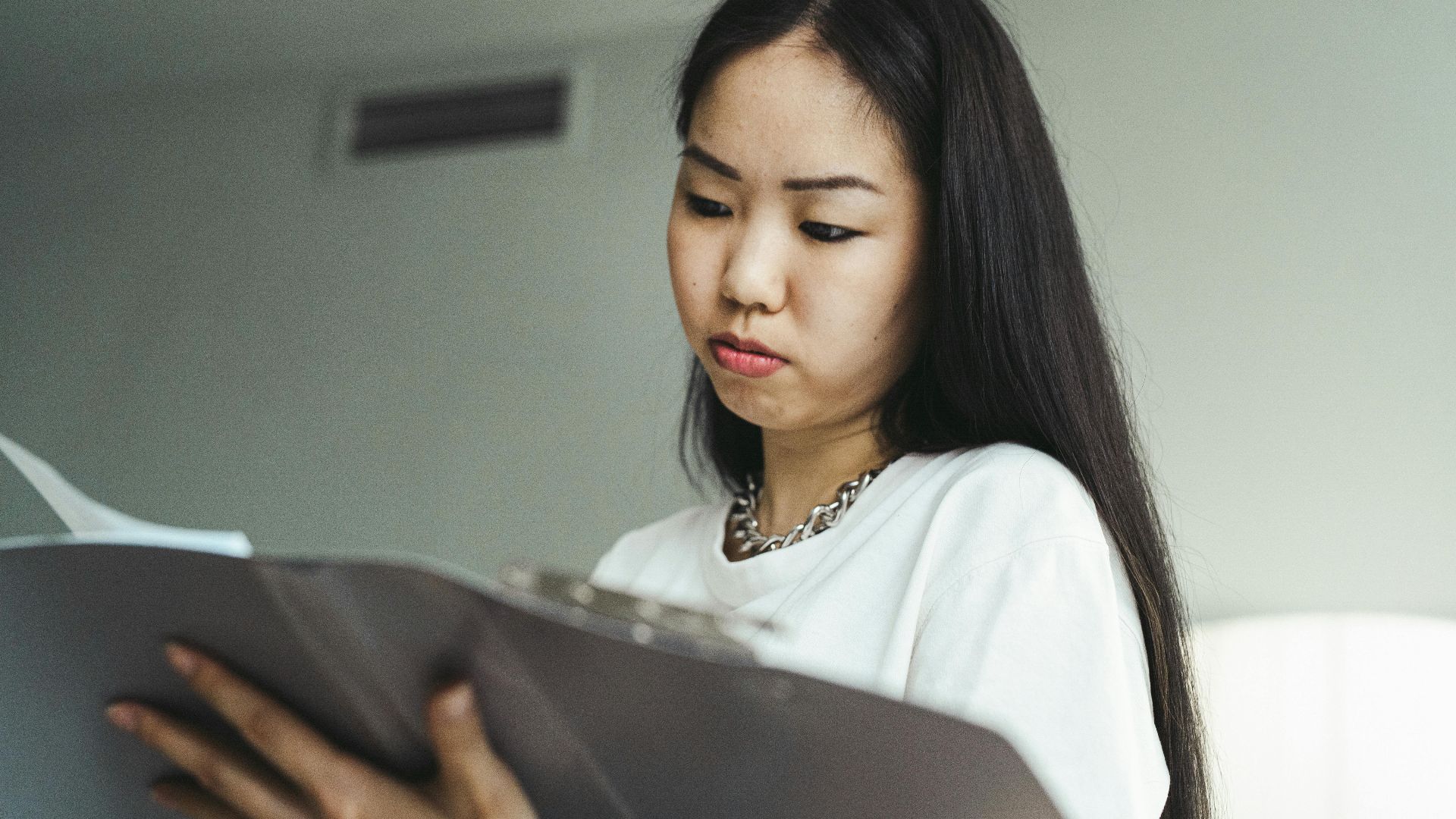Asian woman in casual attire examining paperwork with concentration indoors.
