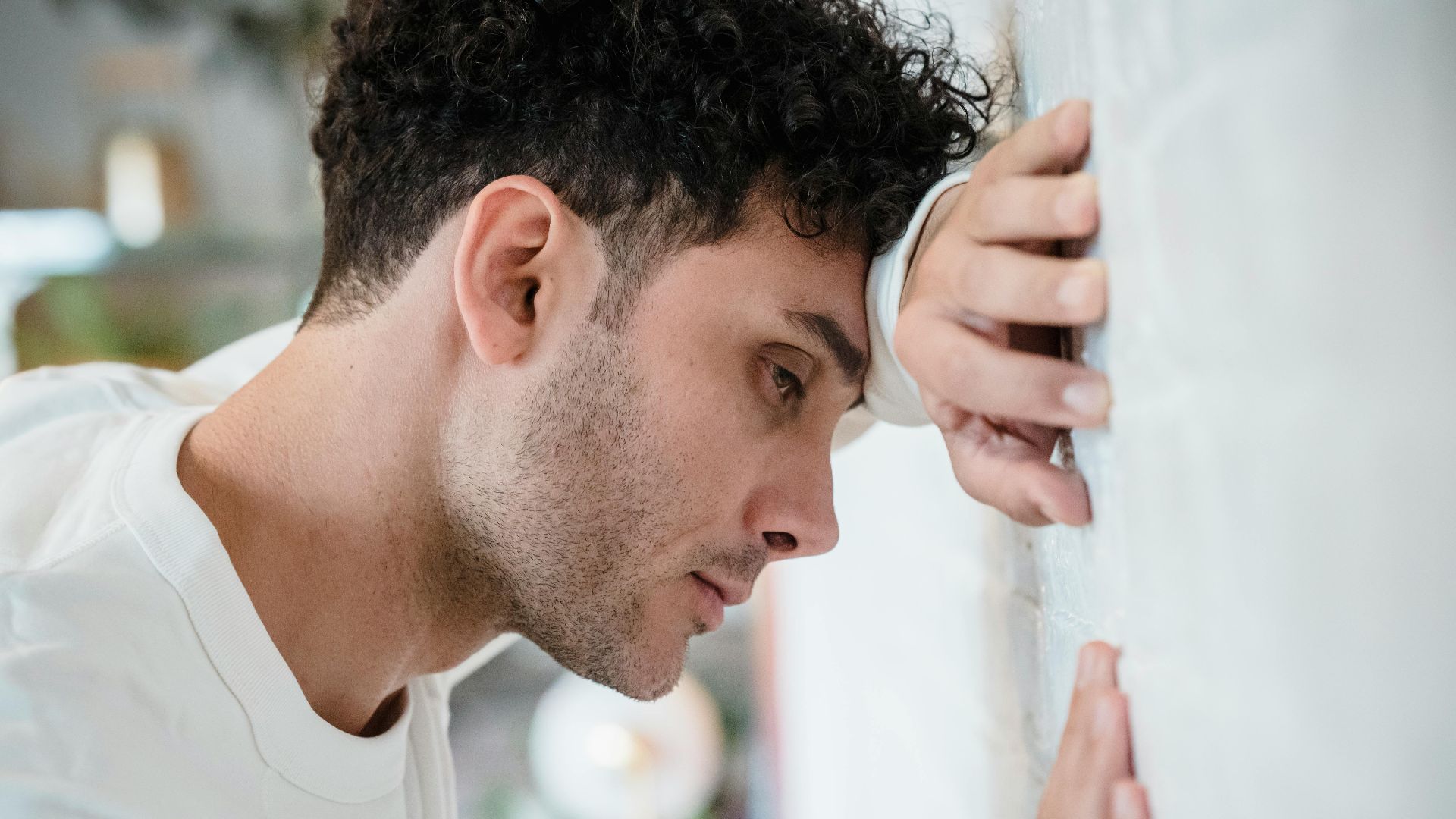 Side view portrait of a man in deep thought leaning against a white wall indoors.