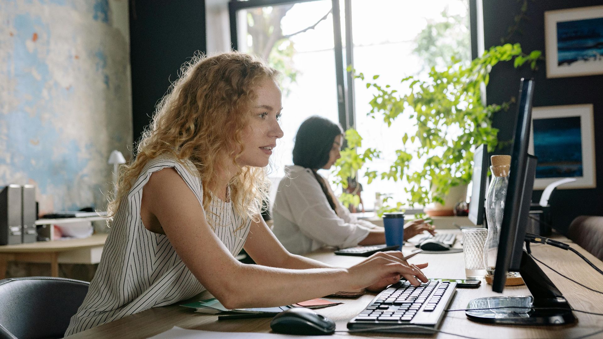 Two focused women working at a bright and modern office space, typing on computers at a shared desk.