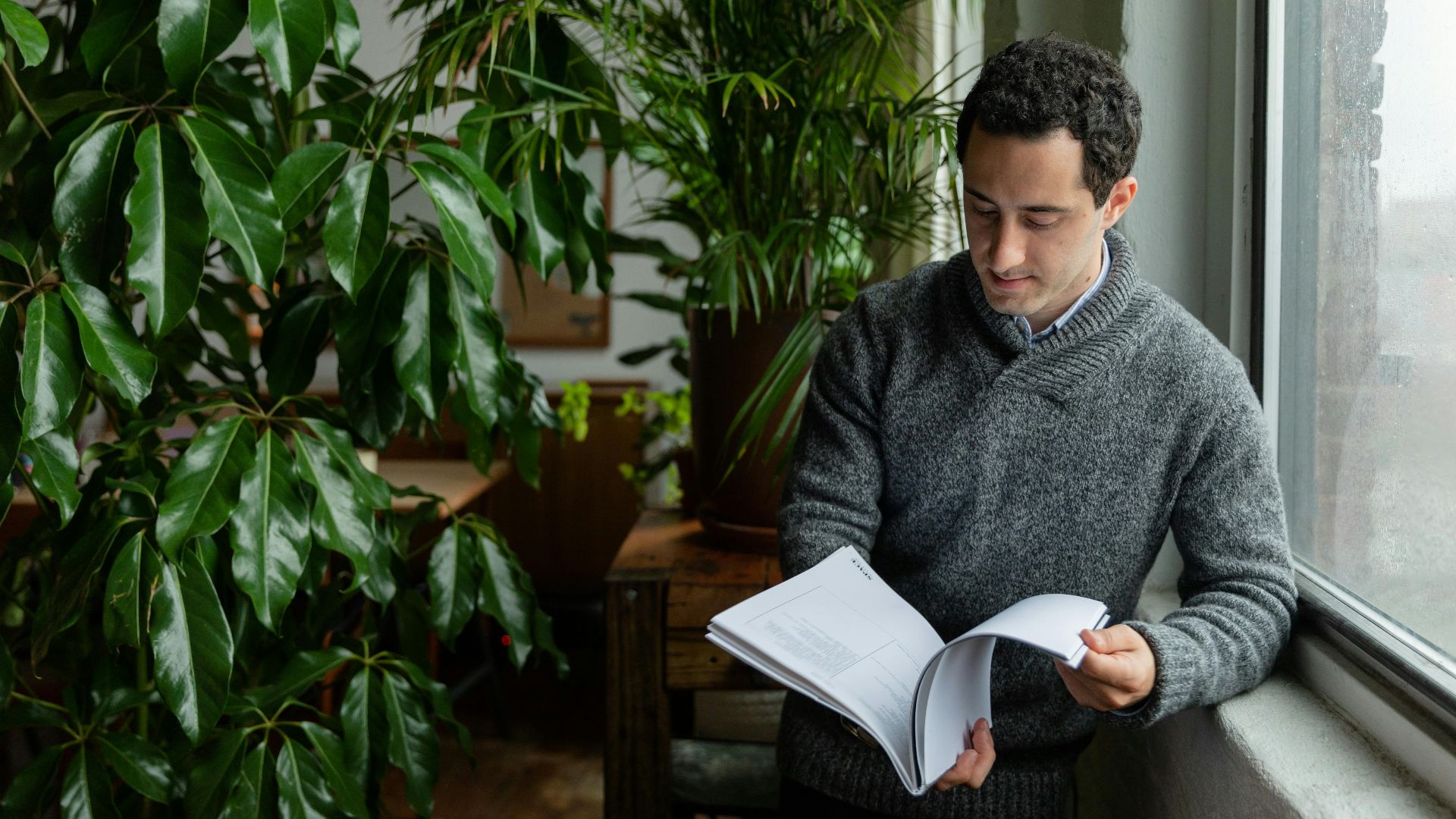 A man in a sweater reads a book next to lush indoor plants by a window.