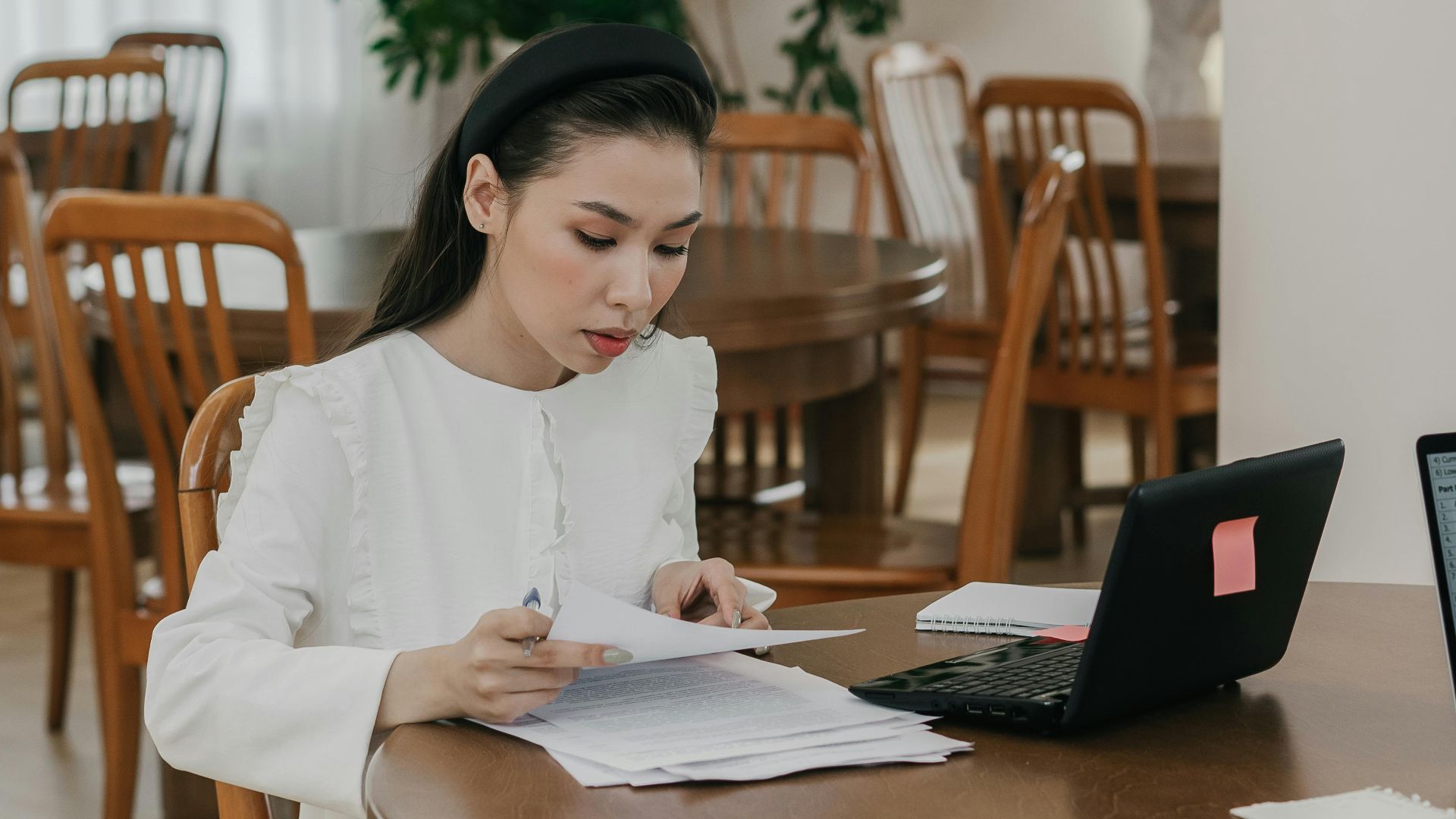 Focused woman studying with papers and laptop in a quiet library setting, surrounded by wooden tables and chairs.