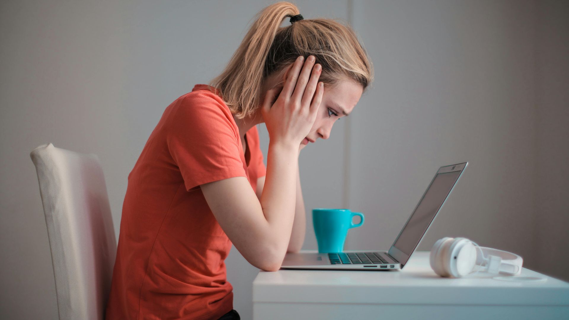 Side view of thoughtful worried female student in casual clothes focusing on screen and trying to solve problem while sitting at table with blue mug of hot drink and headphones and working on laptop at home
