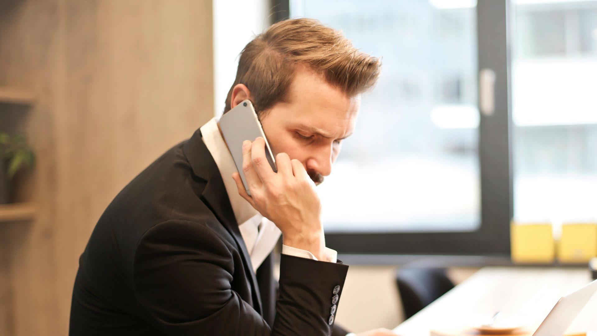 A businessman in a suit making a phone call while working on a laptop in a modern office setting.