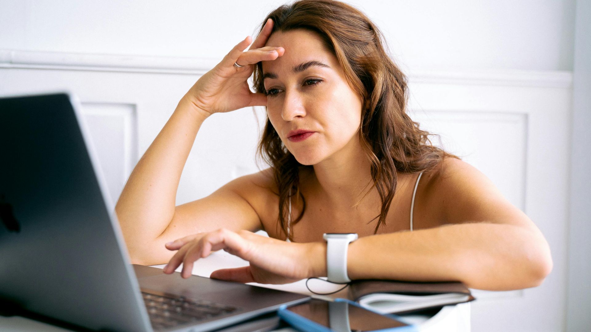 A woman with a frustrated expression, hand on head, working on a laptop.