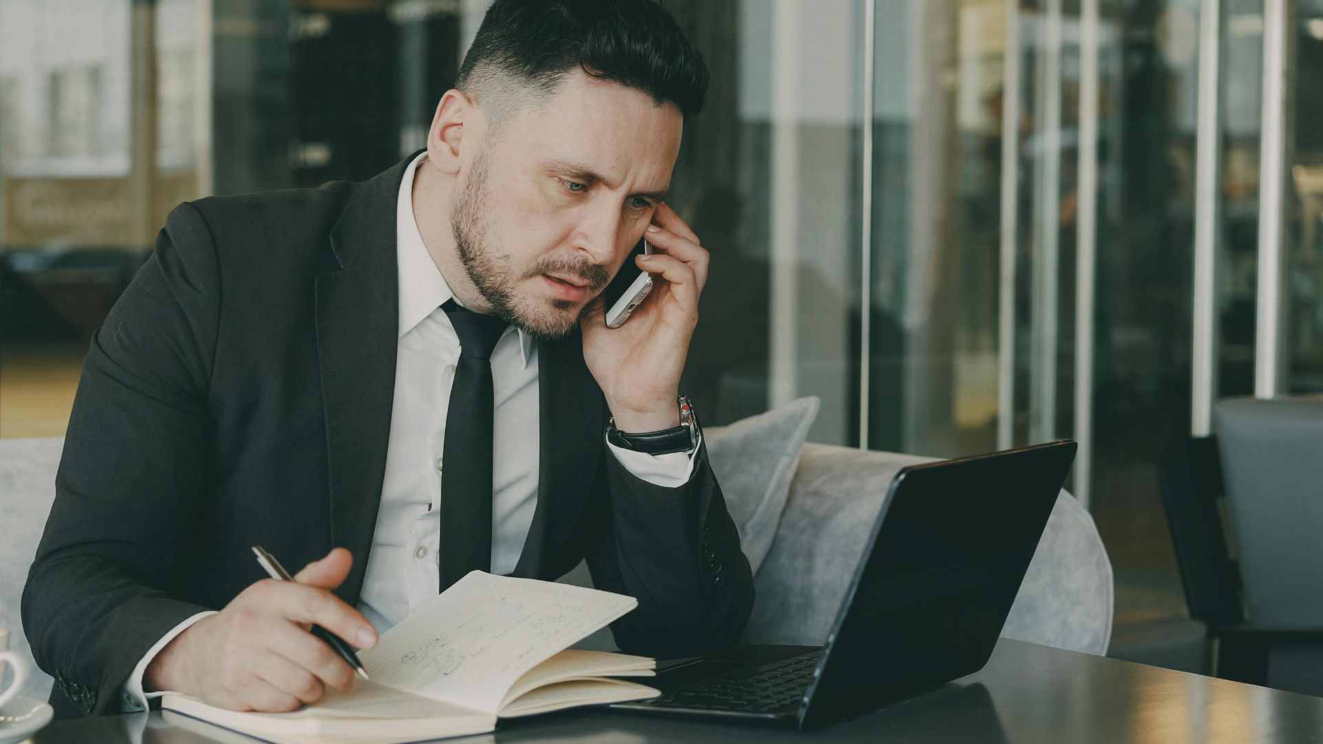 Businessman talks on phone while writing notes in a modern cafe.