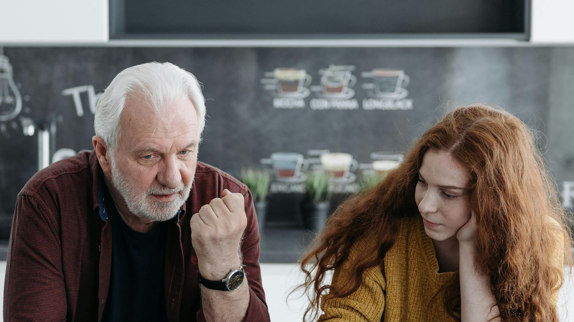 An elderly man and his granddaughter engage in a heartfelt conversation over coffee indoors.