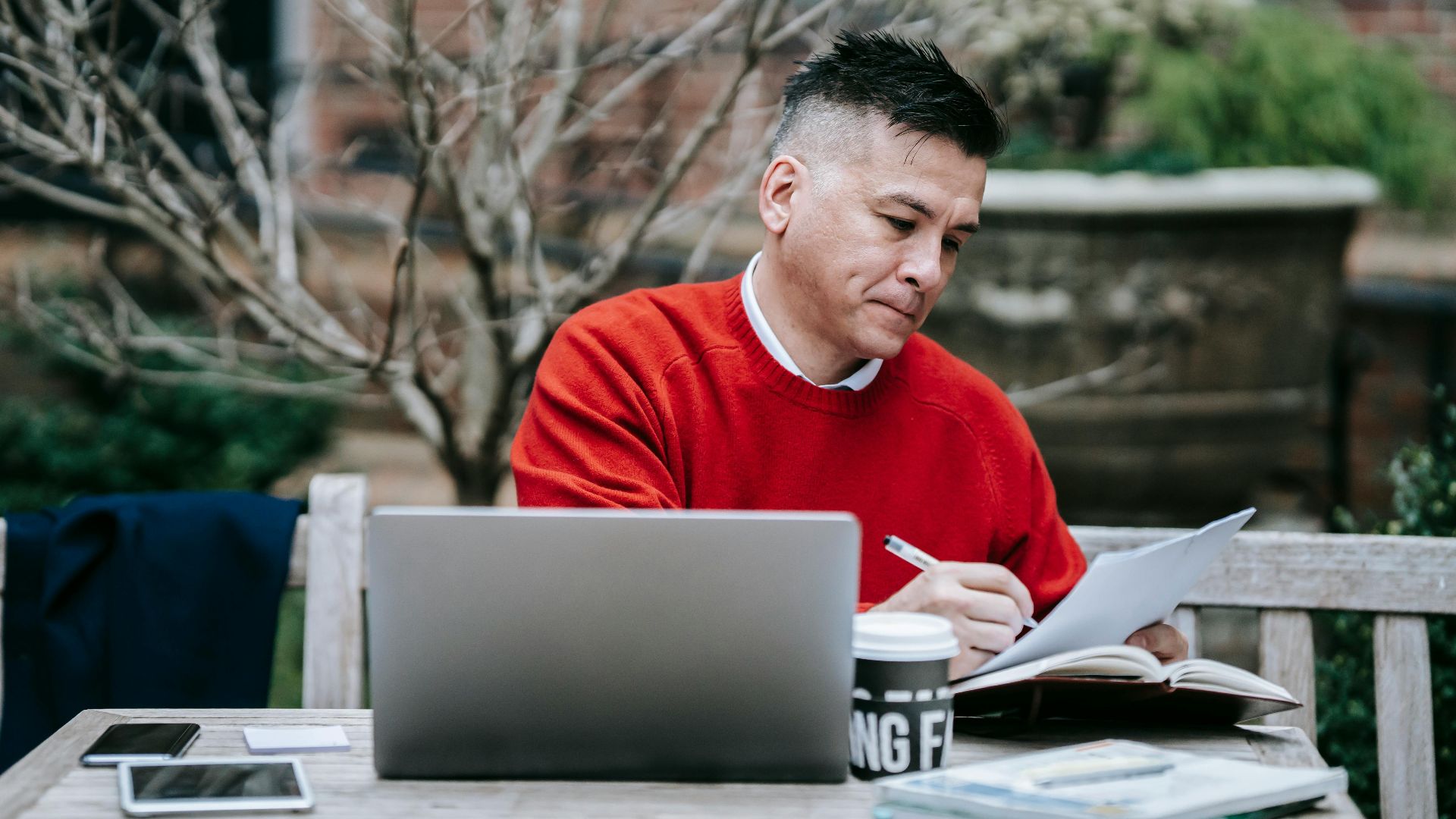 A man in a red sweater working on paperwork with a laptop outdoors on a wooden bench.