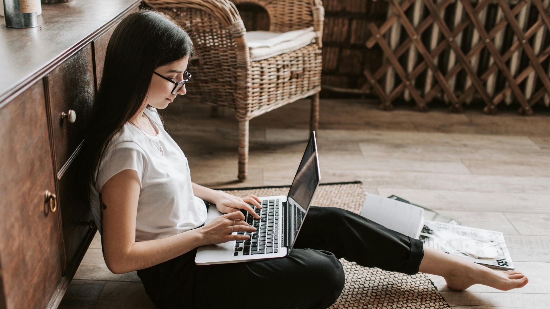 Side view of happy young woman with long dark hair in eyeglasses typing on keyboard of computer while sitting on floor in modern living room