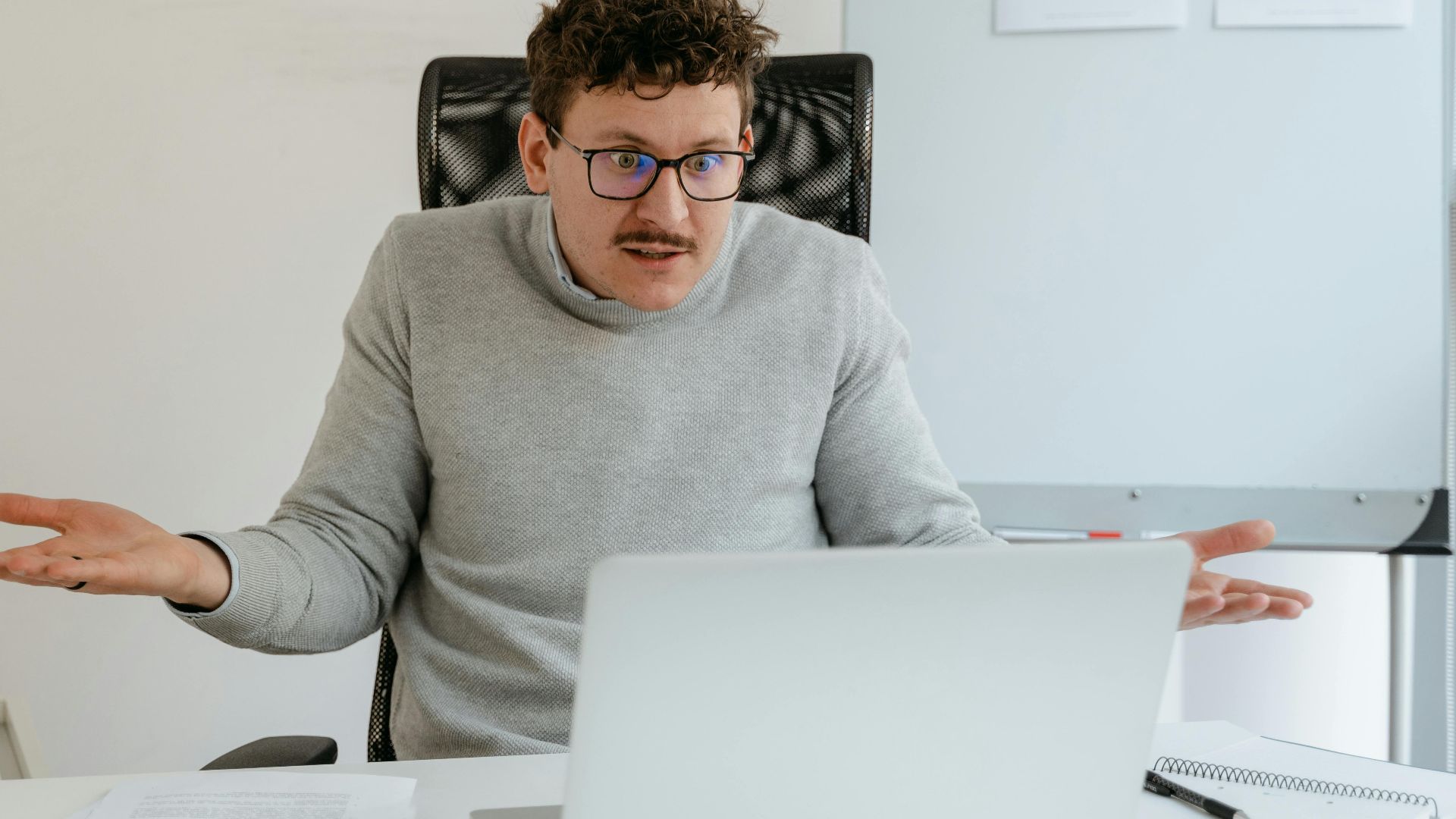 A man in a gray sweater and glasses gesturing at his laptop screen during a video call in an office setting.