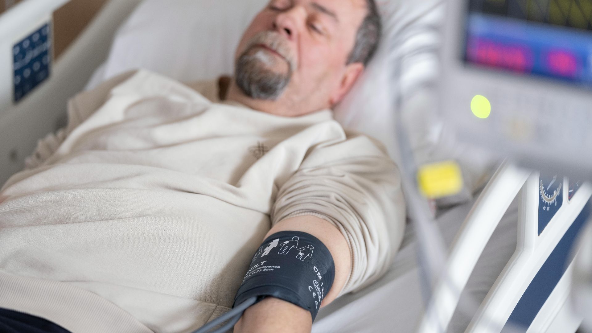 Elderly man resting in hospital bed with blood pressure monitor attached, symbolizing healthcare and medical care.