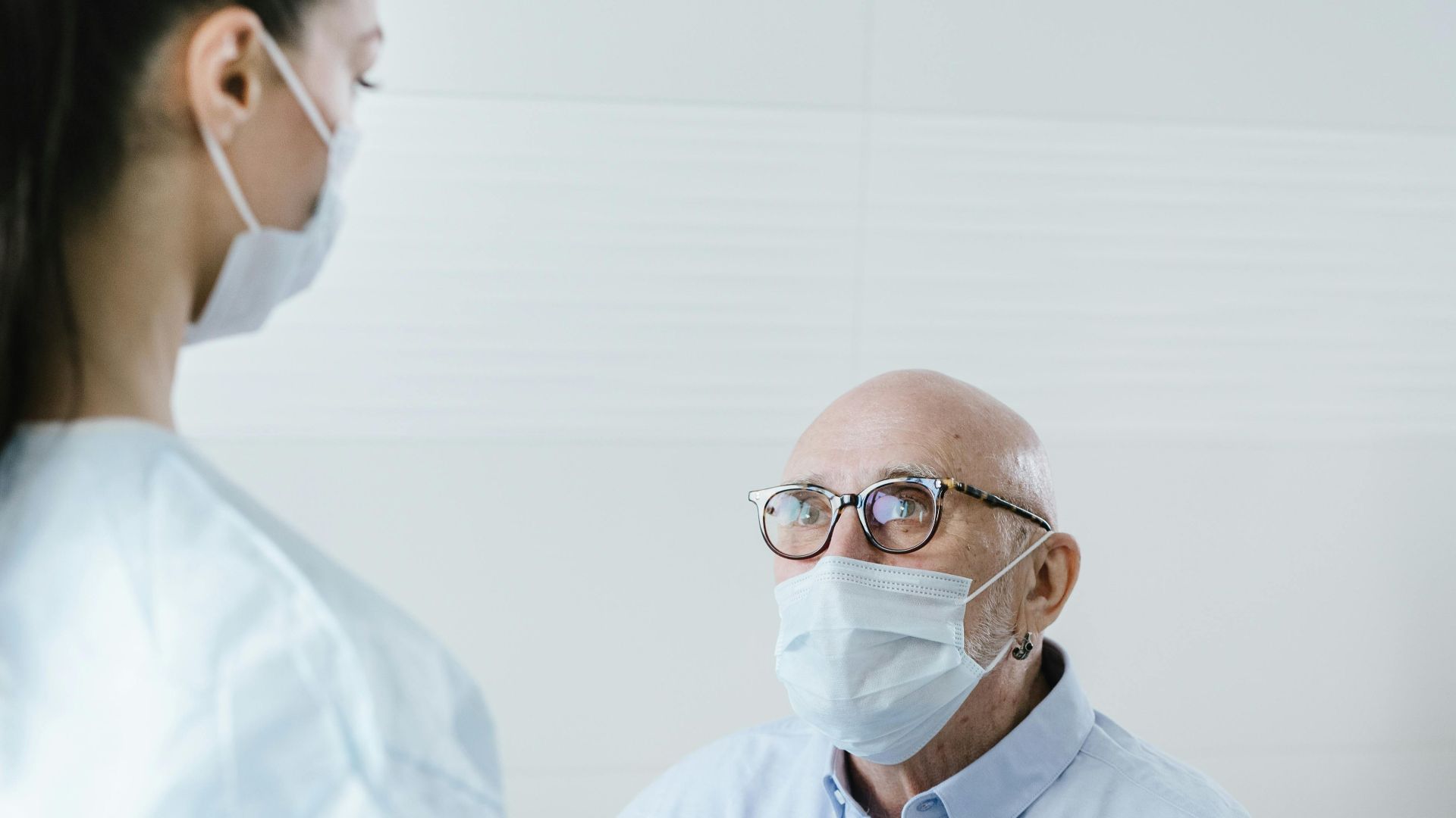 A medical professional discusses health with a senior patient, both wearing face masks for safety.