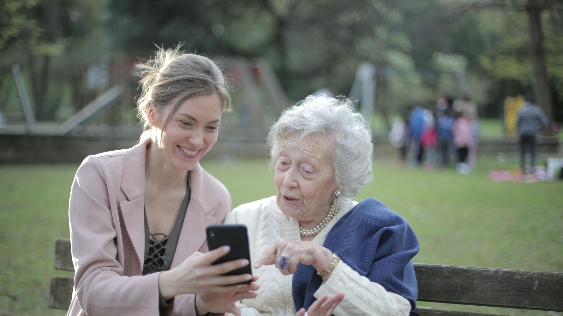 Delighted female relatives sitting together on wooden bench in park and browsing mobile phone while learning using