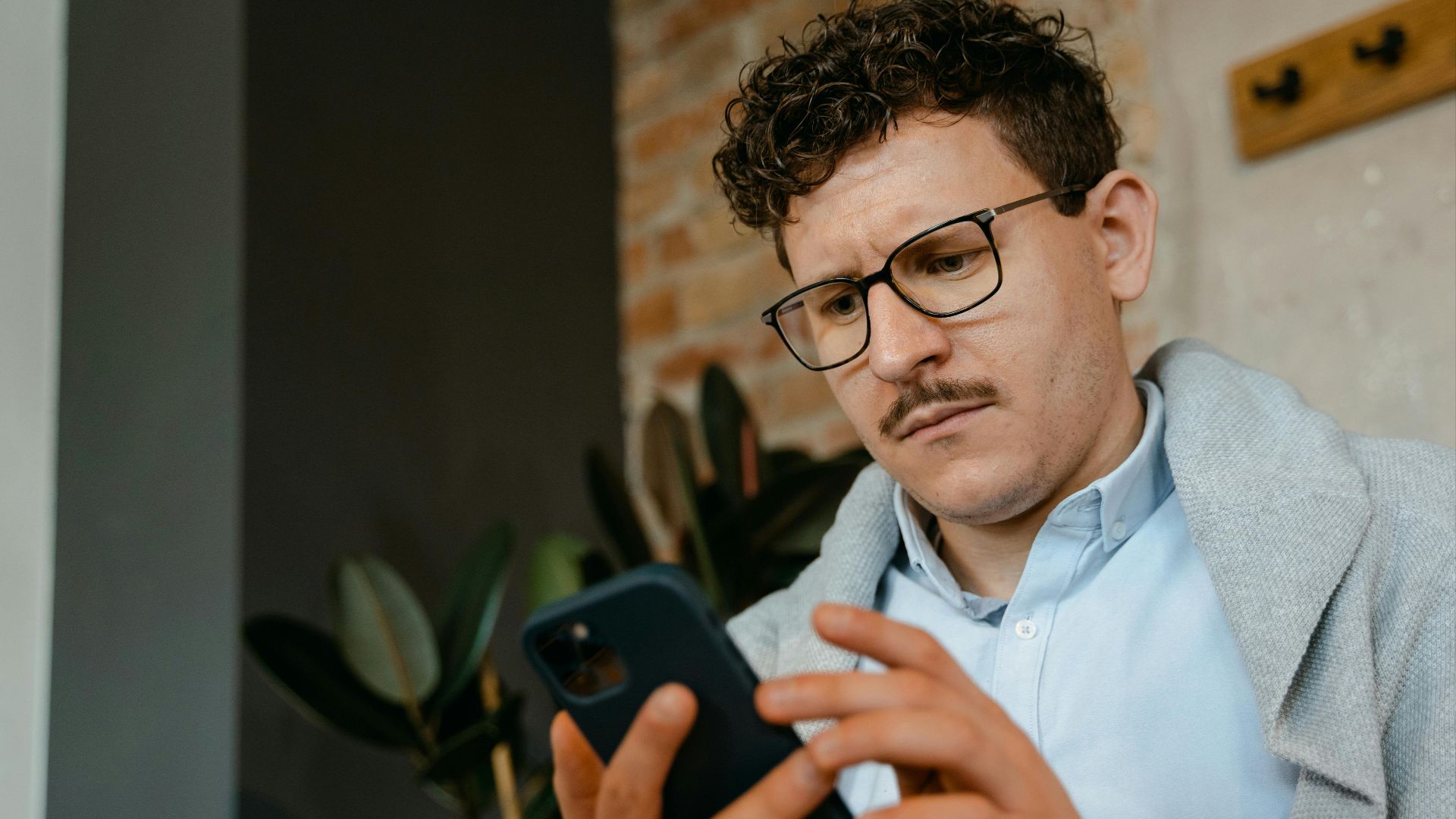 A focused man with glasses using a smartphone indoors next to a brick wall.