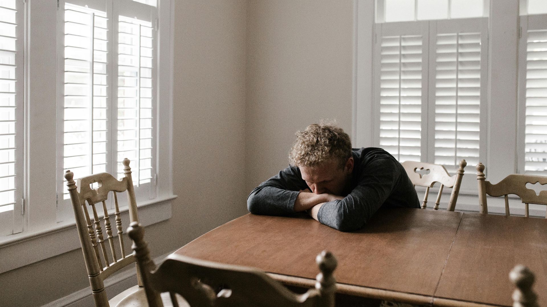 An adult man with emotions of sadness and anxiety sitting alone at a wooden table near windows.