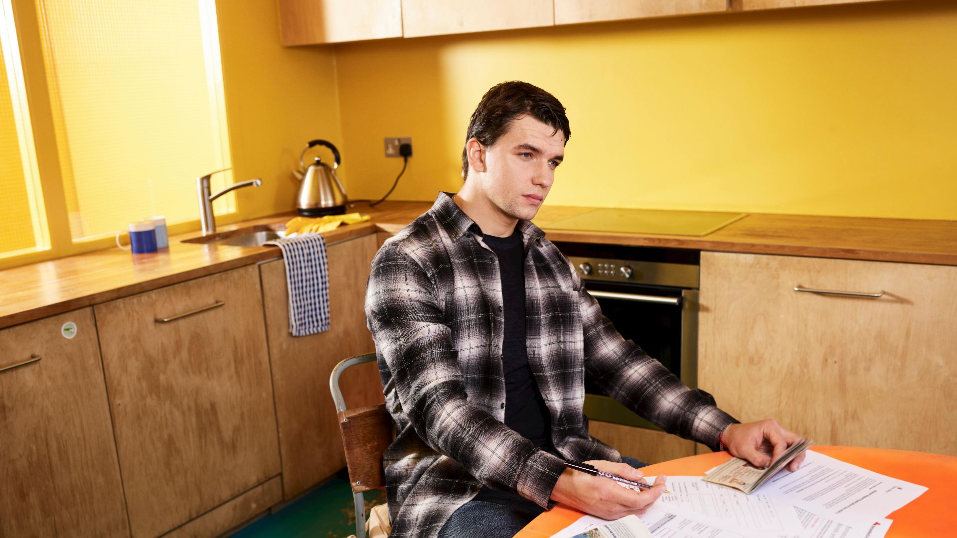 Man sitting in kitchen reviewing bills and a check with smartphone nearby.