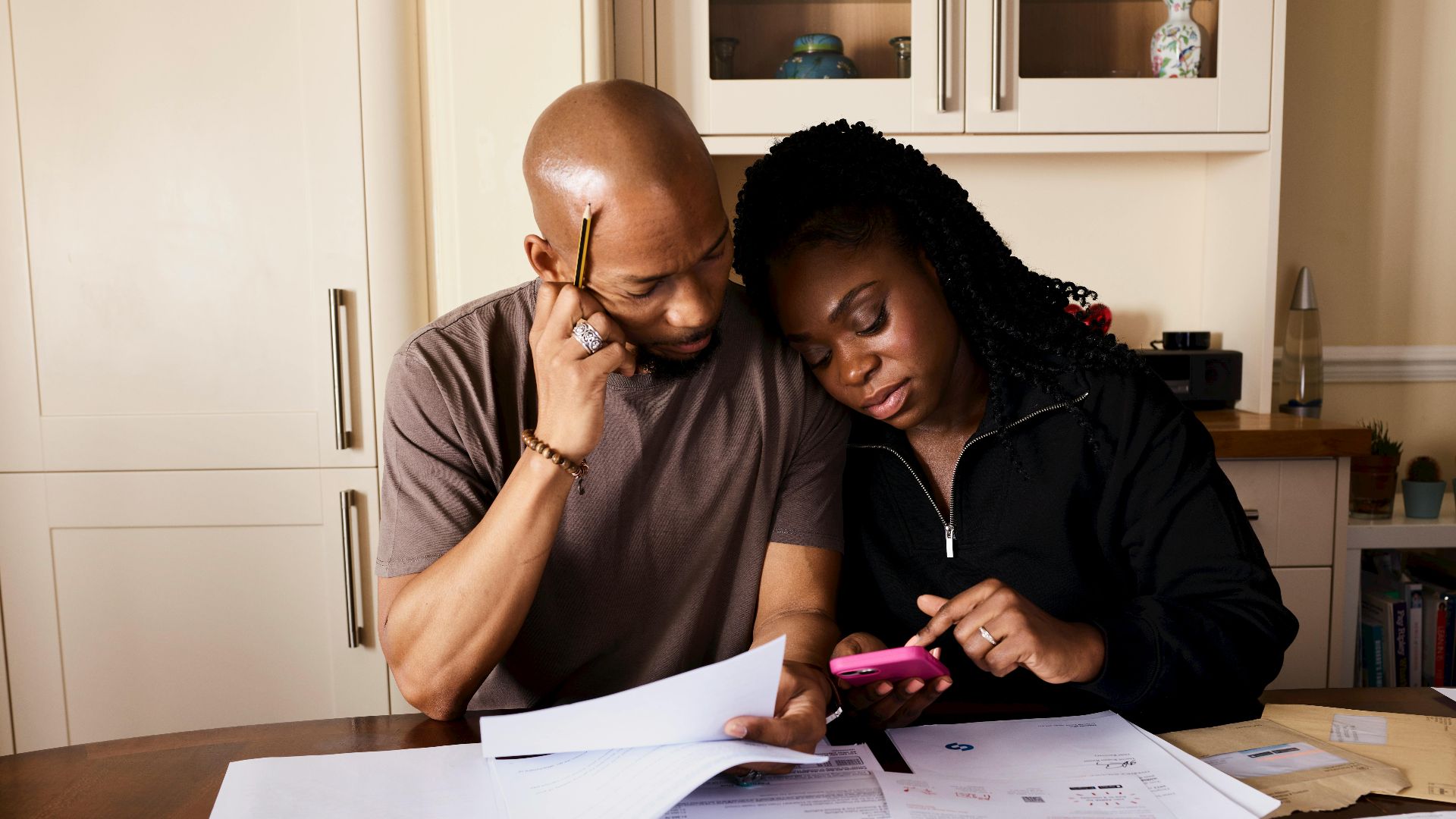 A couple sits at a table managing domestic finances, evaluating documents and using a smartphone.