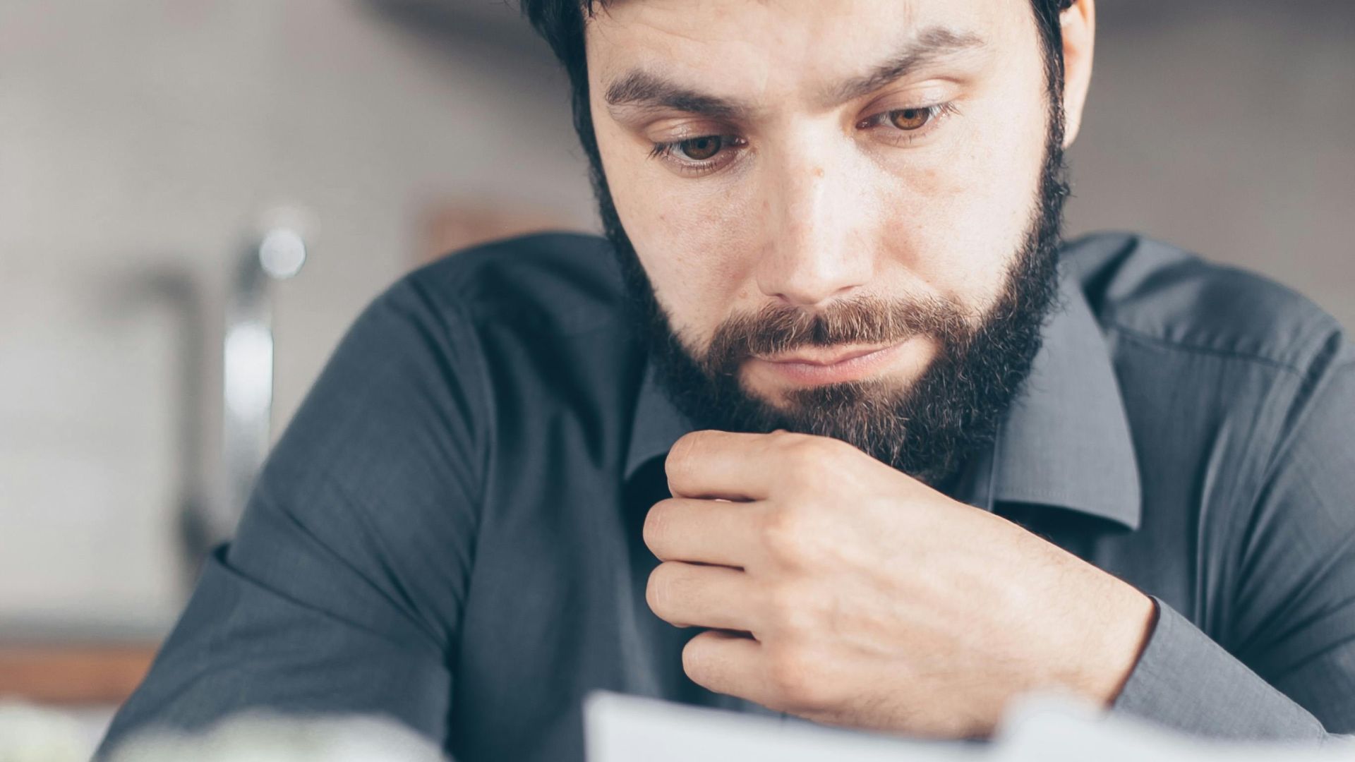 A bearded man in a gray shirt intently reads documents while seated indoors, appearing concerned.