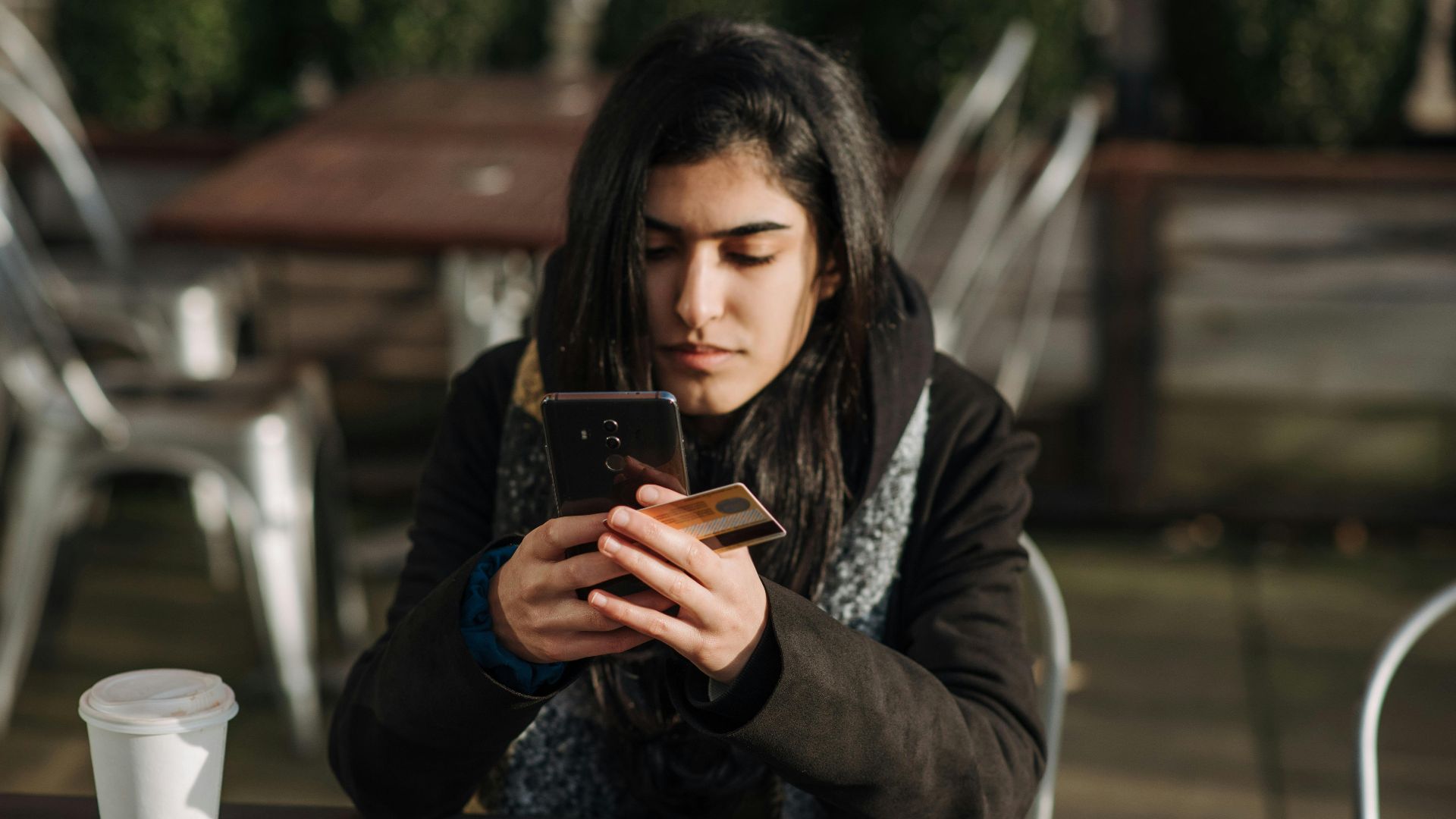 Young ethnic female purchaser with credit card shopping online on mobile phone at table with takeaway hot drink in cafeteria