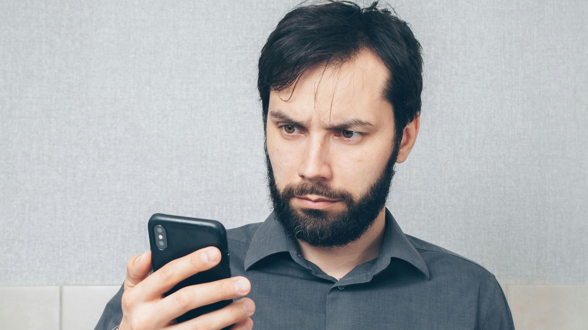 An adult man in a gray shirt looks concerned while using a smartphone and holding a credit card indoors.