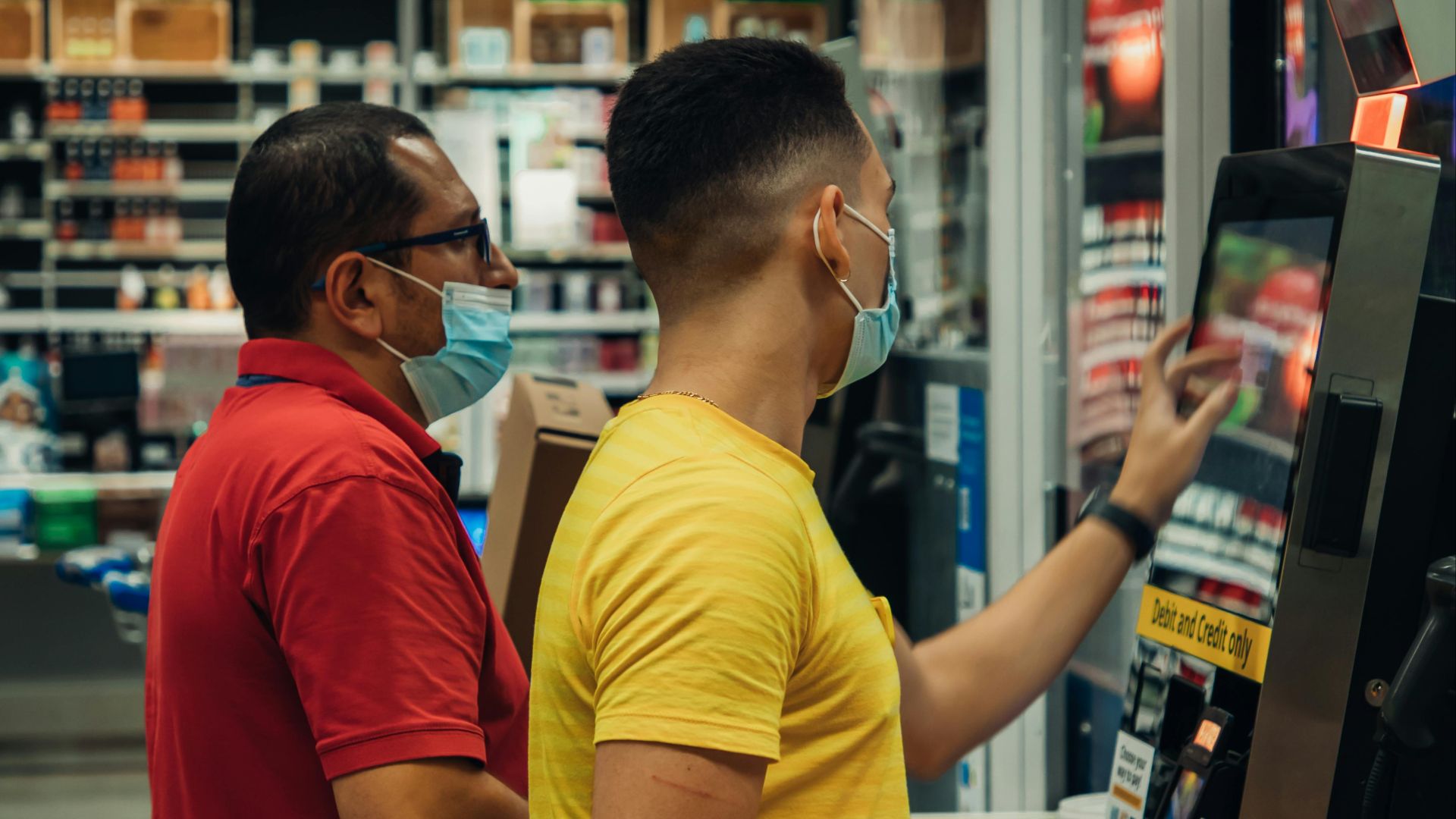 Two men wearing masks using a self-checkout machine in a grocery store.