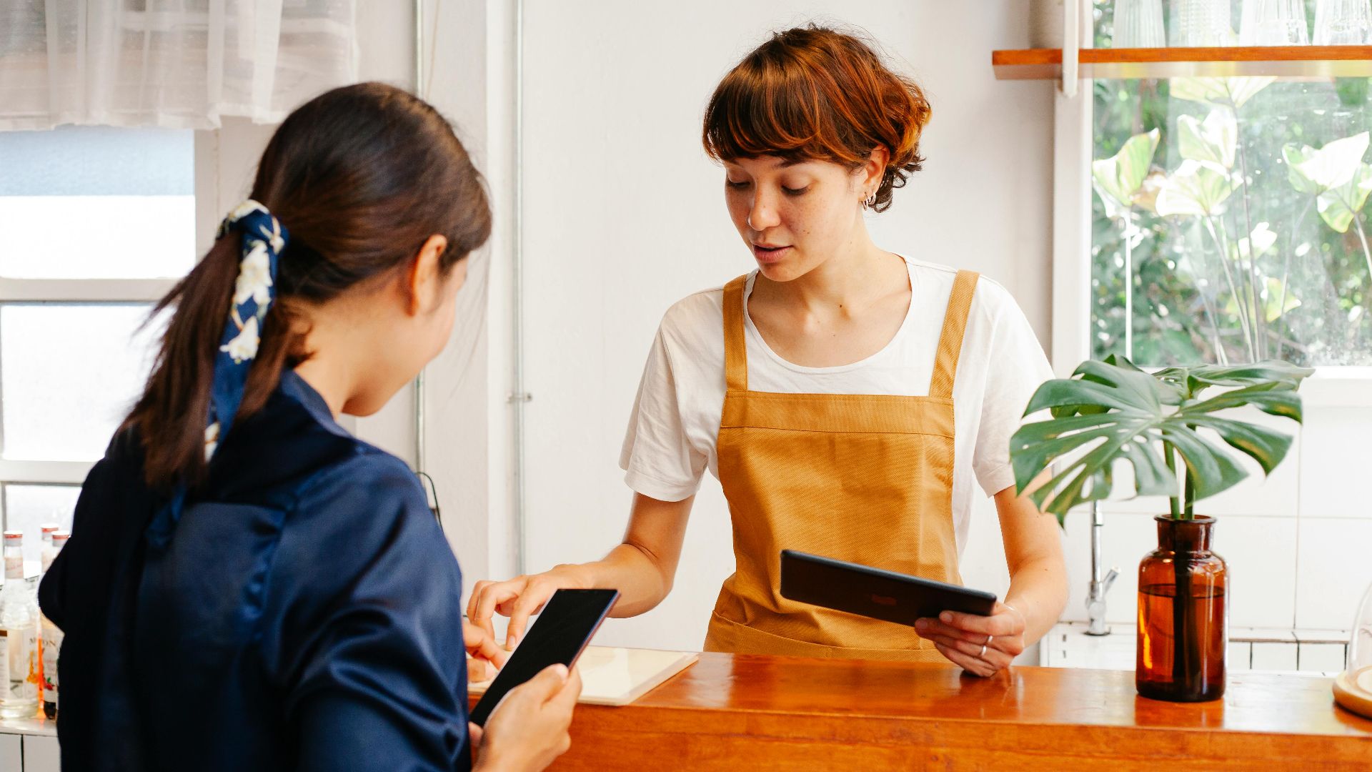 Unrecognizable female cafeteria worker with cellphone talking to young colleague with tablet in daylight
