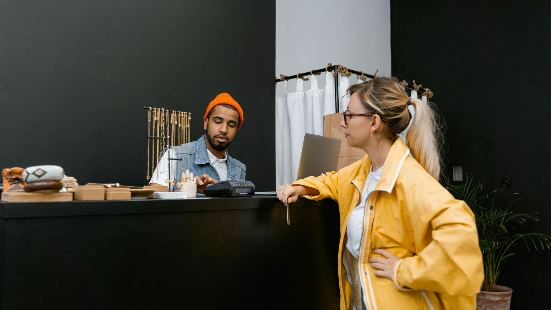 A woman pays with a card at a store counter to a bearded cashier in a denim vest.