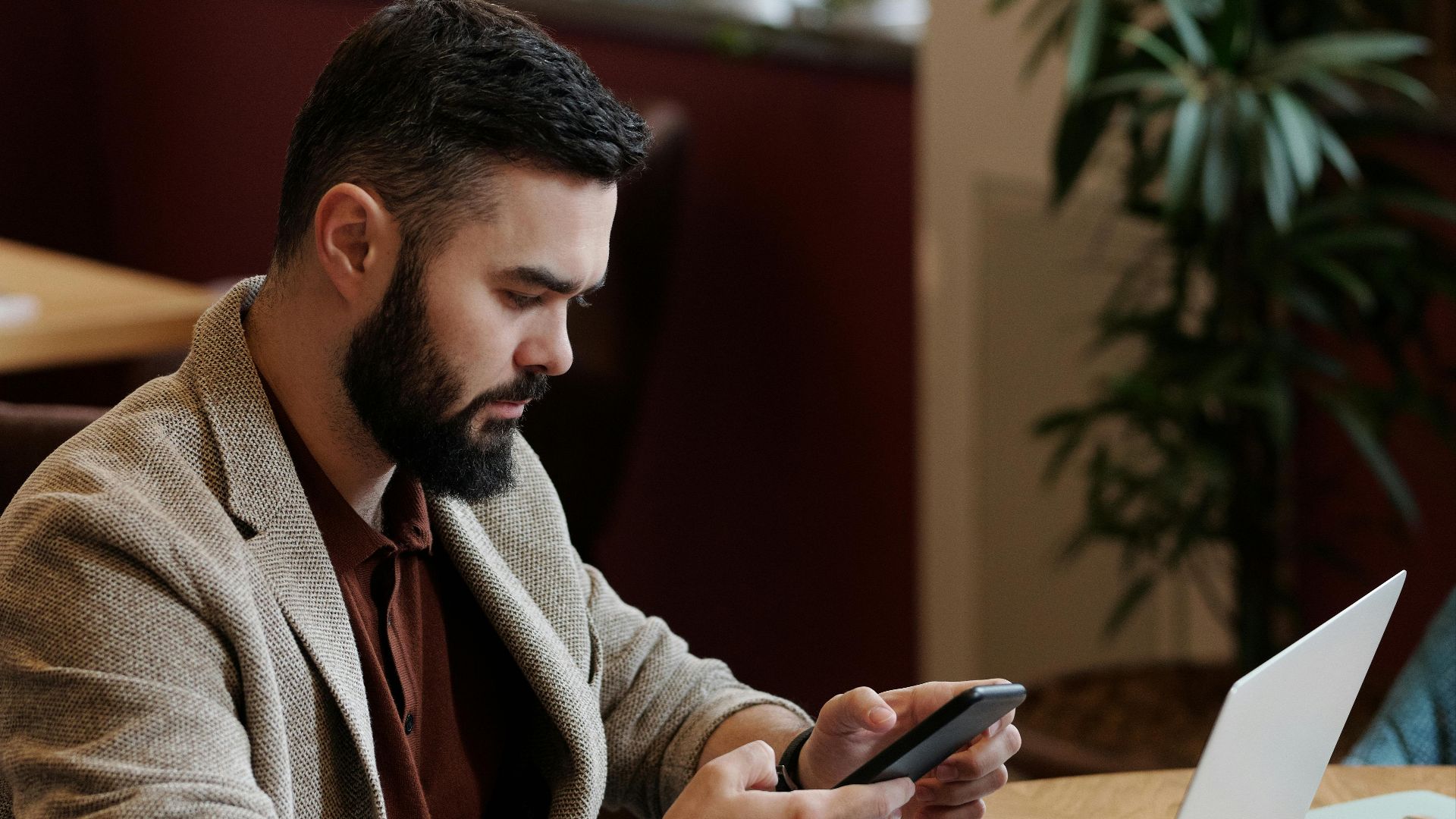 Bearded man using smartphone and laptop at café with coffee, focused on remote work.