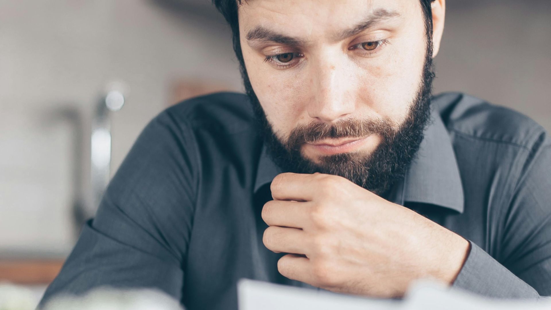 A bearded man in a gray shirt intently reads documents while seated indoors, appearing concerned.