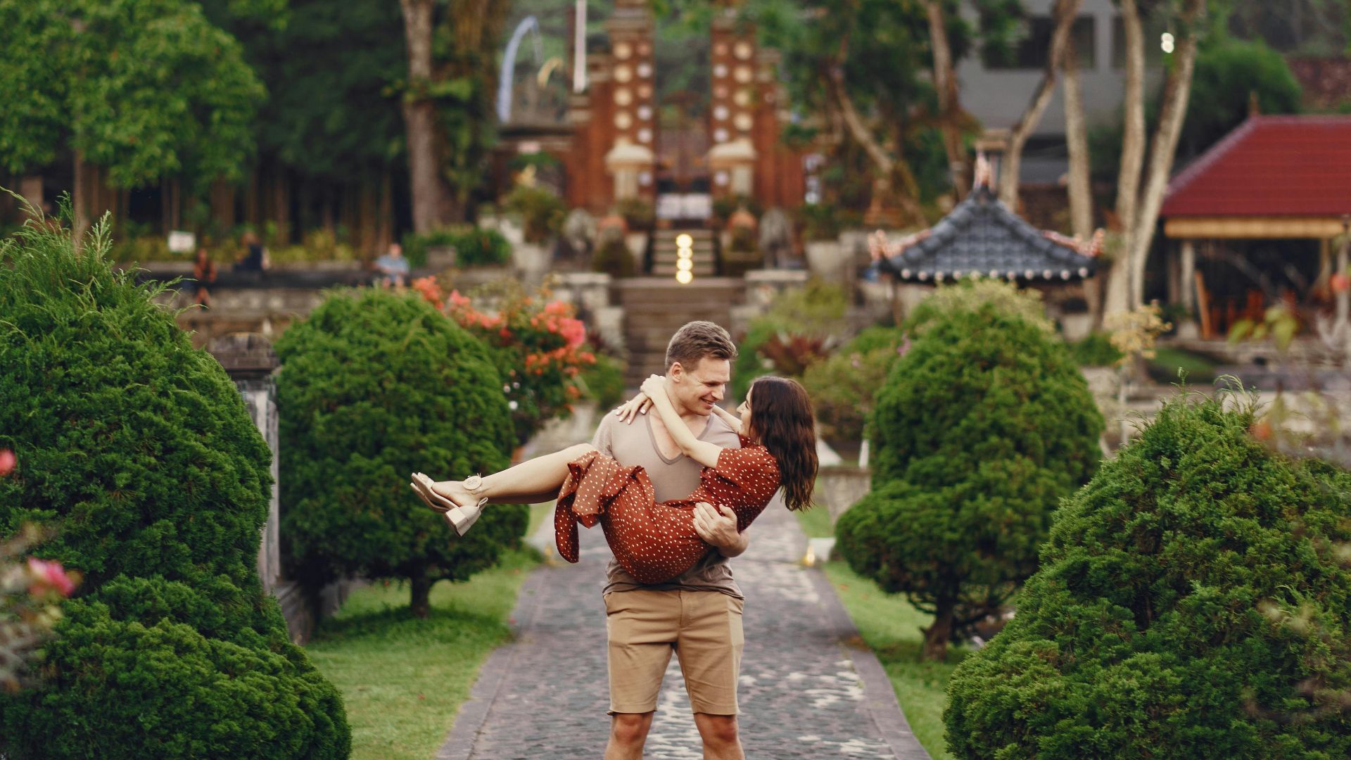 Happy young man in casual outfit carrying woman in arms on alley in tropical park while sightseeing during romantic journey