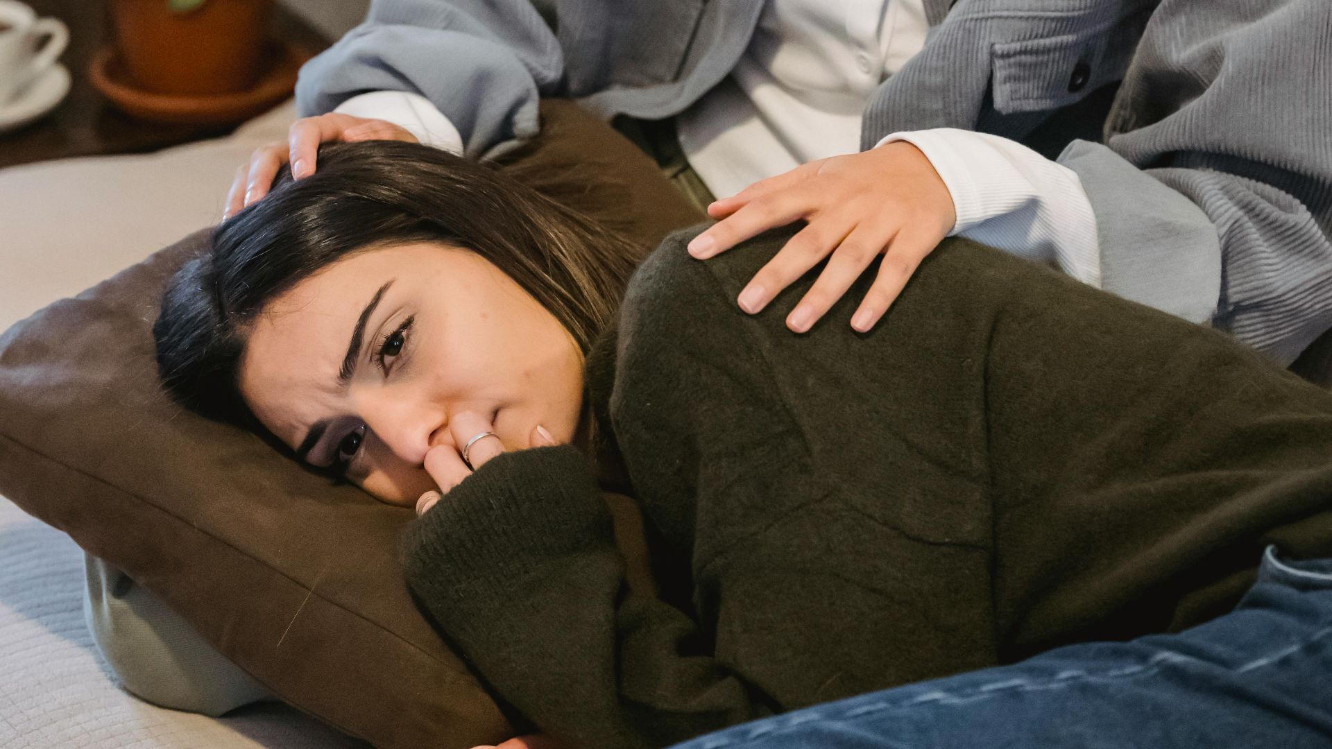 Calm young ethnic lady with curly hair sitting on sofa and calming sad crying female friend lying on knees