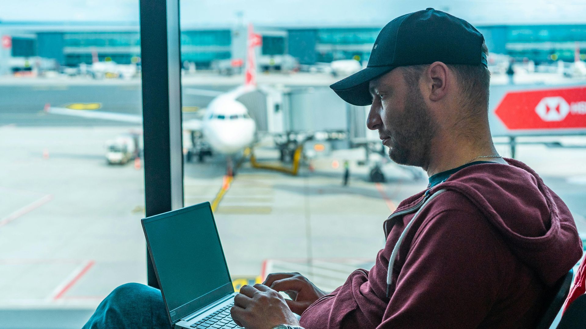 A man working on his laptop in an airport terminal, with a plane visible outside.