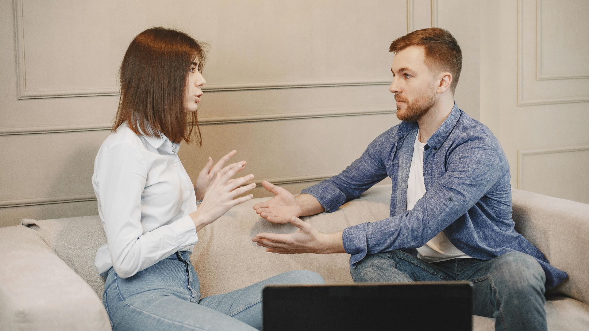 A young couple sitting on a couch having a thoughtful conversation with expressive hand gestures.