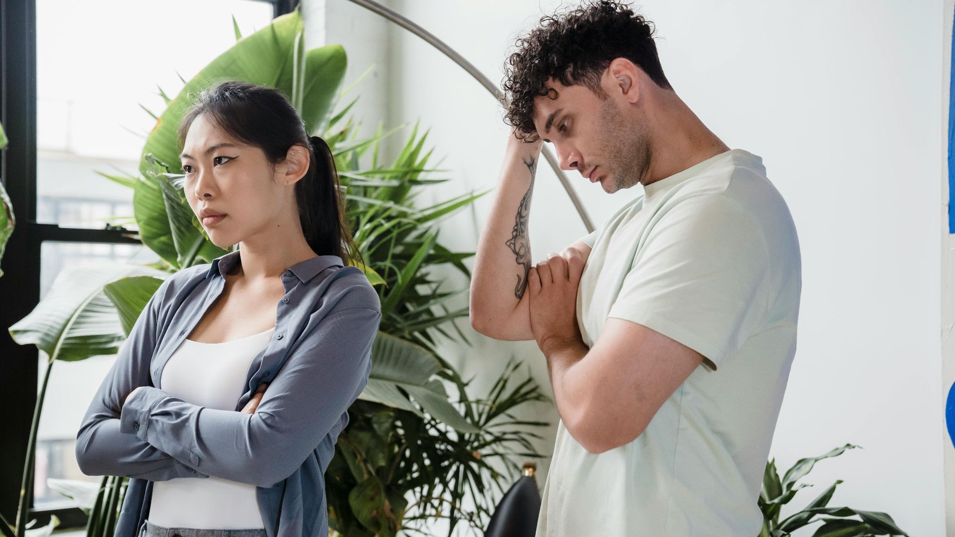 A man and woman stand indoors appearing in conflict, surrounded by lush plants.