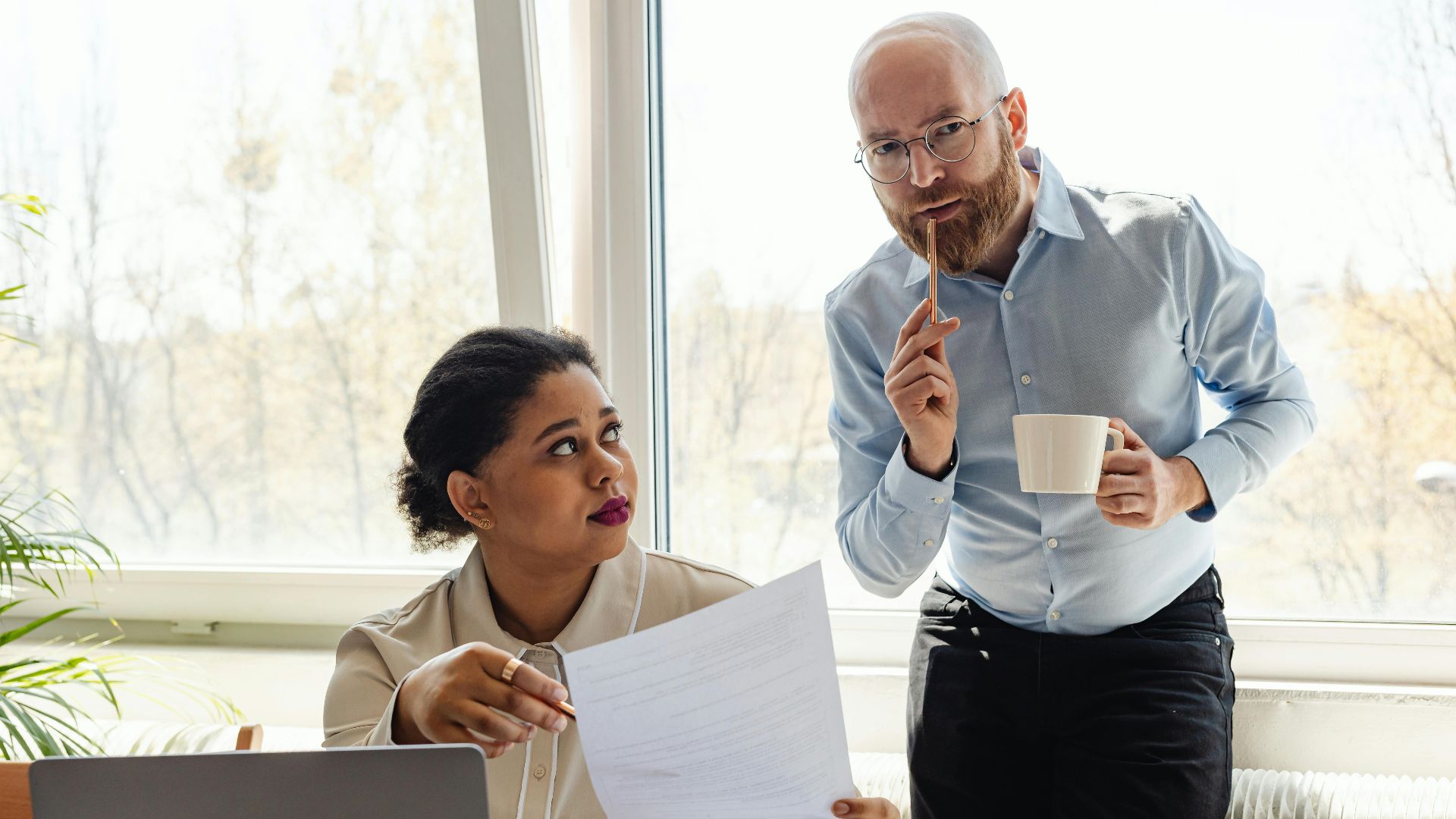 Two colleagues discussing work with laptop and documents in a bright office setting.