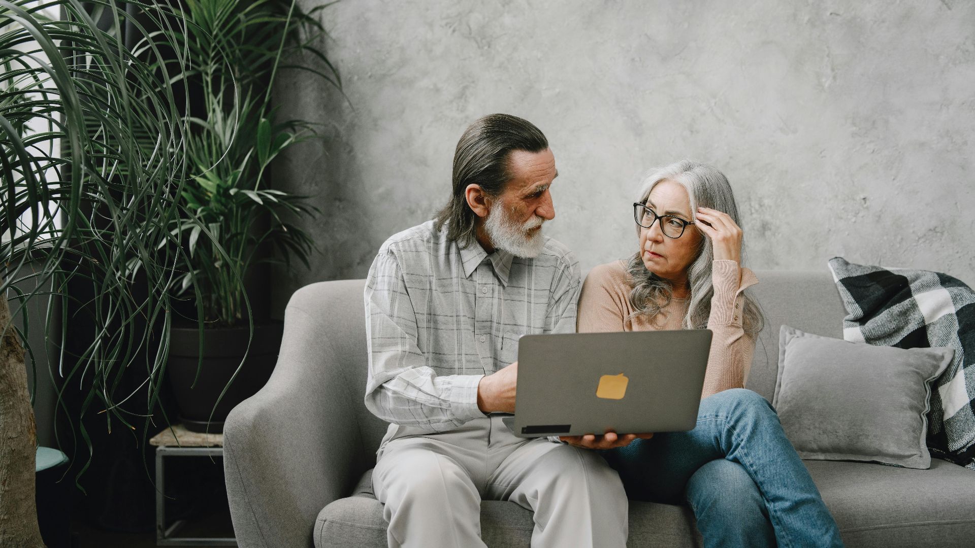 Elderly couple discussing while using laptop at home on sofa, surrounded by plants.