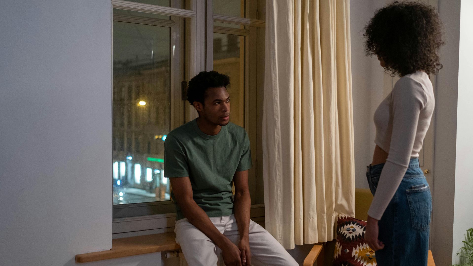 A couple engaged in a tense conversation by a windowsill in a cozy living room setting.