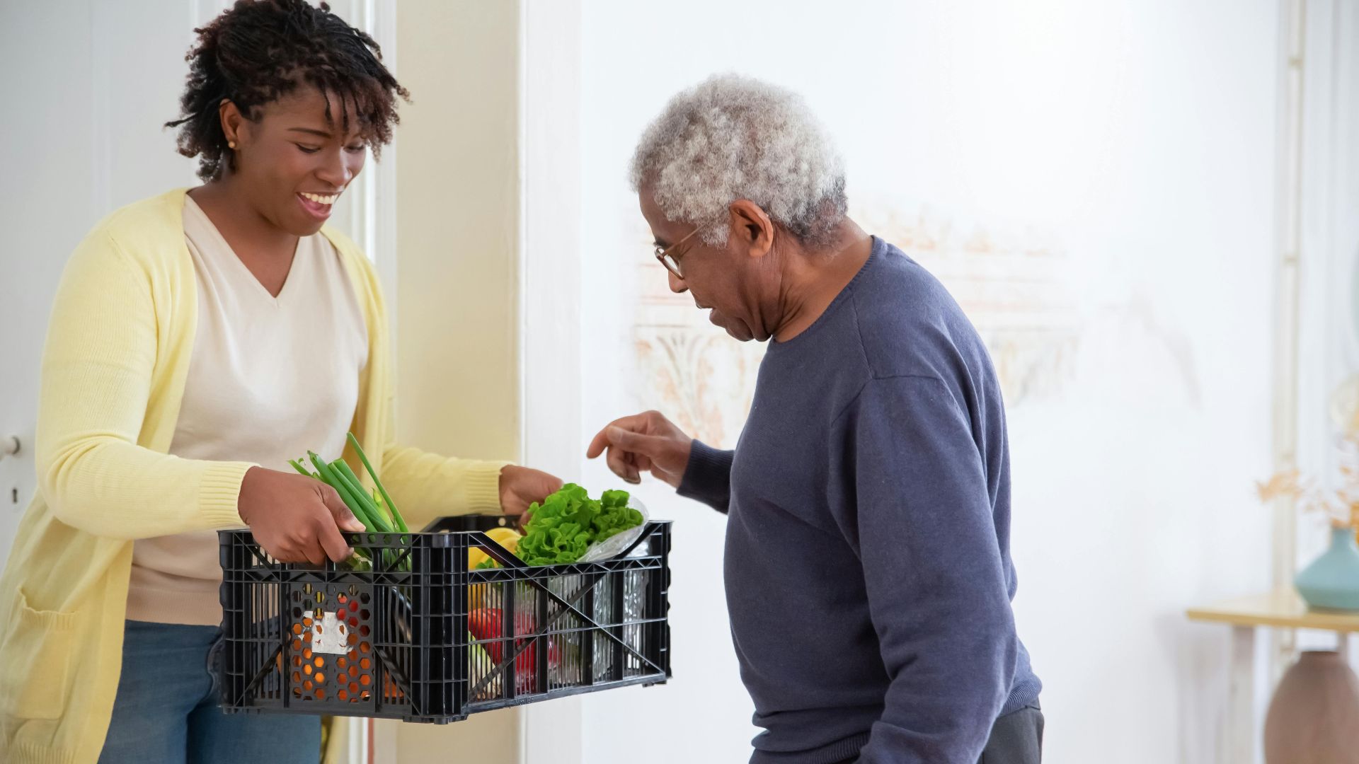 A young adult woman helps a senior man by carrying a crate of fresh vegetables indoors, symbolizing care and support.