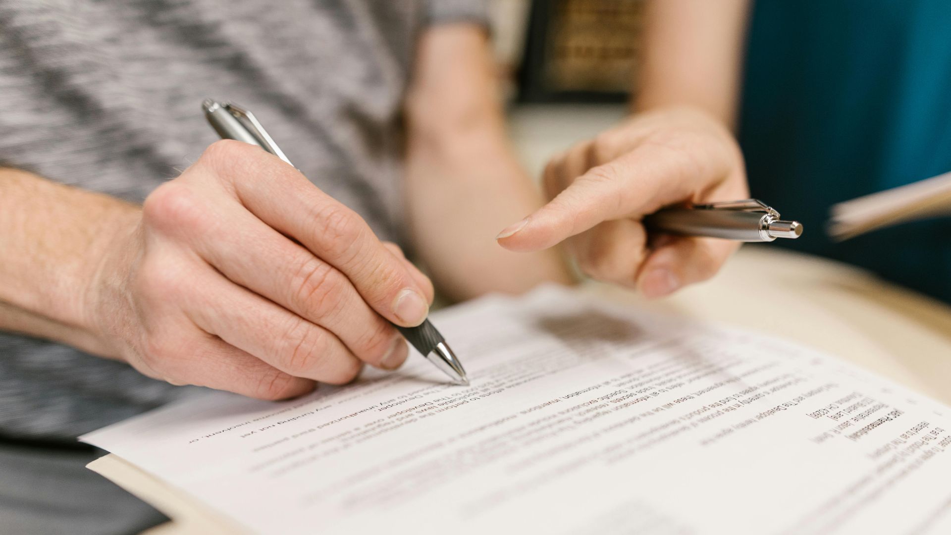 Close-up of two adults reviewing and signing a legal document with pens indoors.