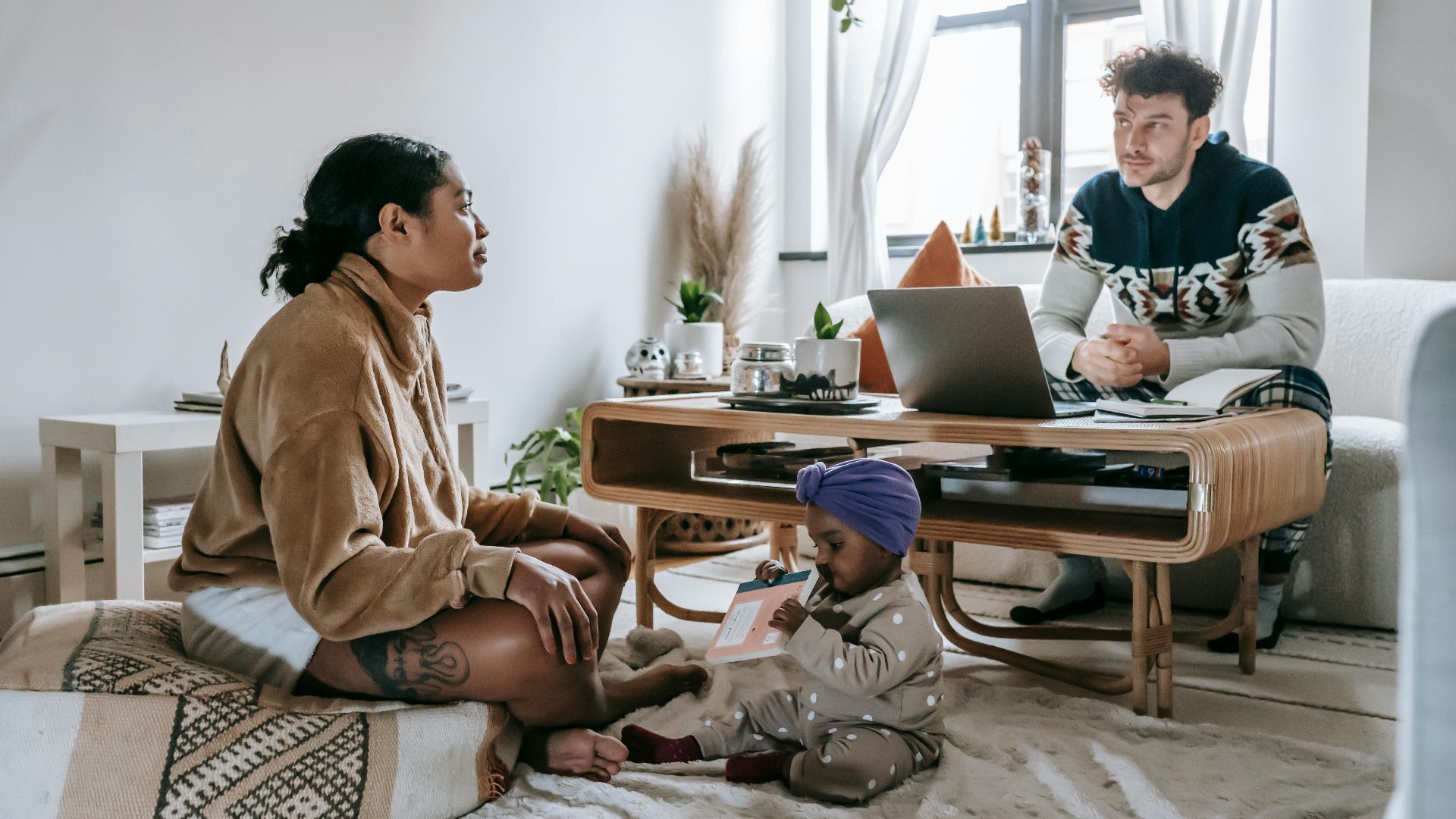 Full body of serious ethnic man sitting near computer in living room with black woman and little toddler at home