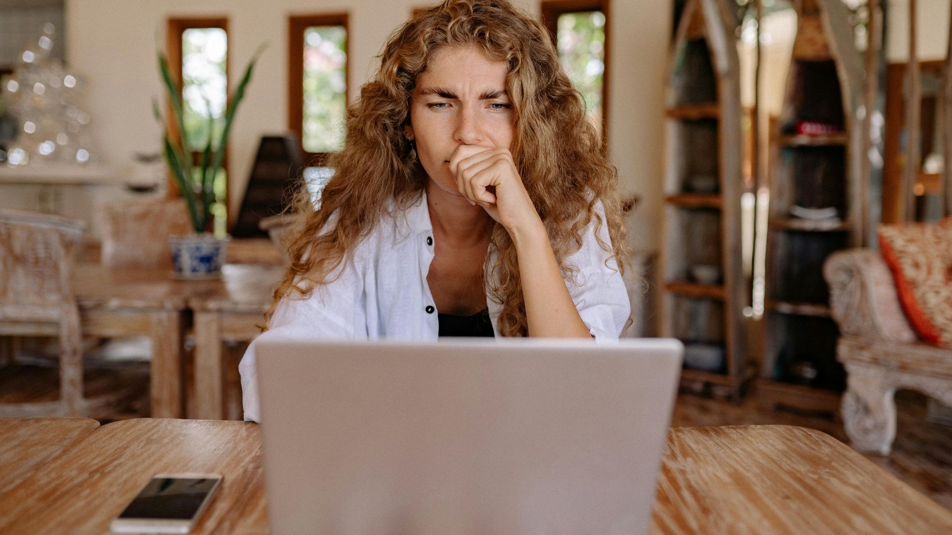 A woman working on her laptop at home, deeply contemplating her work, symbolizing remote work challenges.