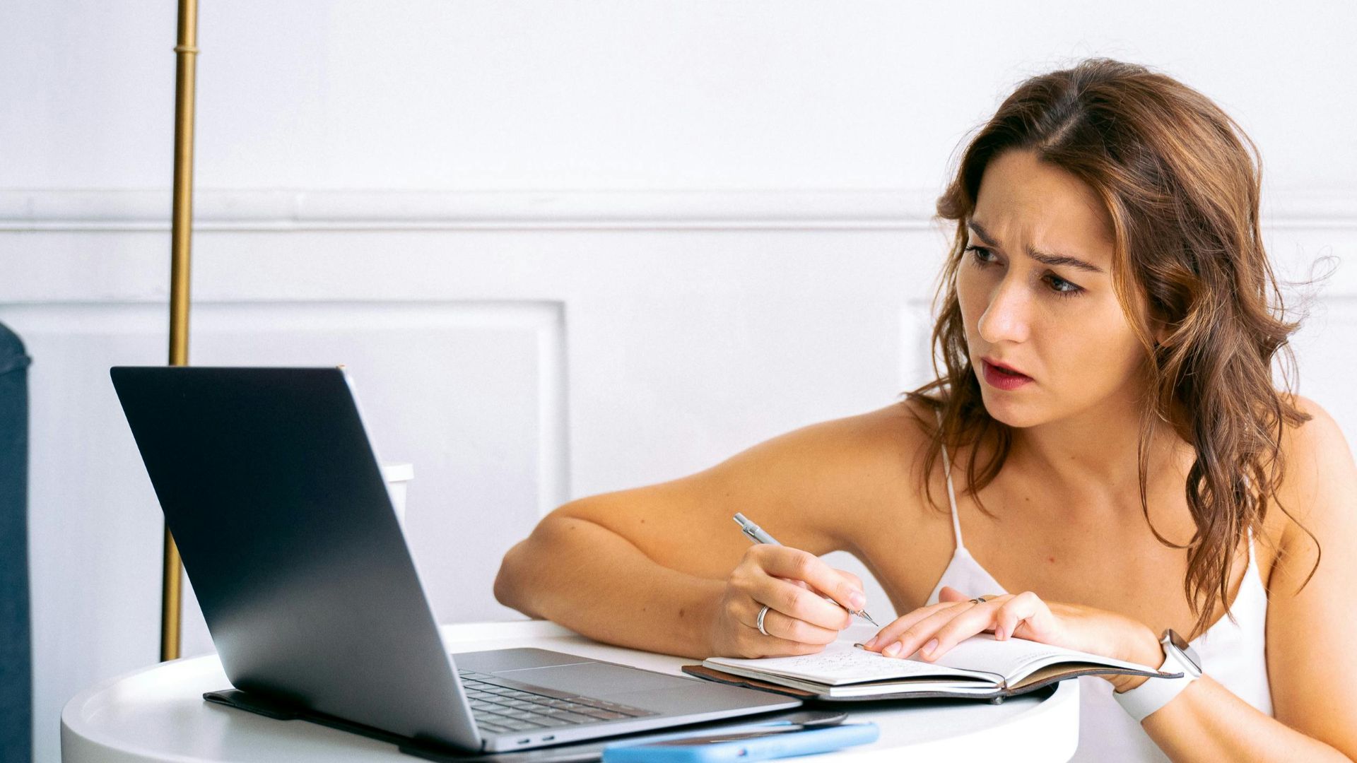 A woman in a white top attentively takes notes using a laptop at home.