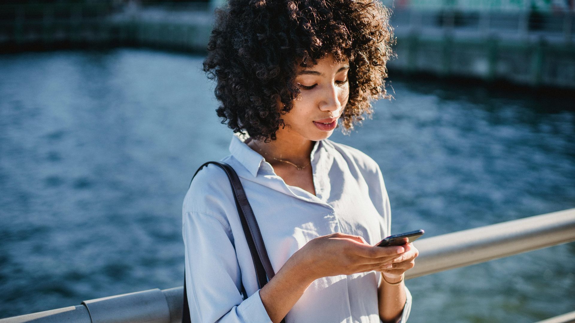 Young woman using her smartphone on a sunny day by the waterfront bridge.