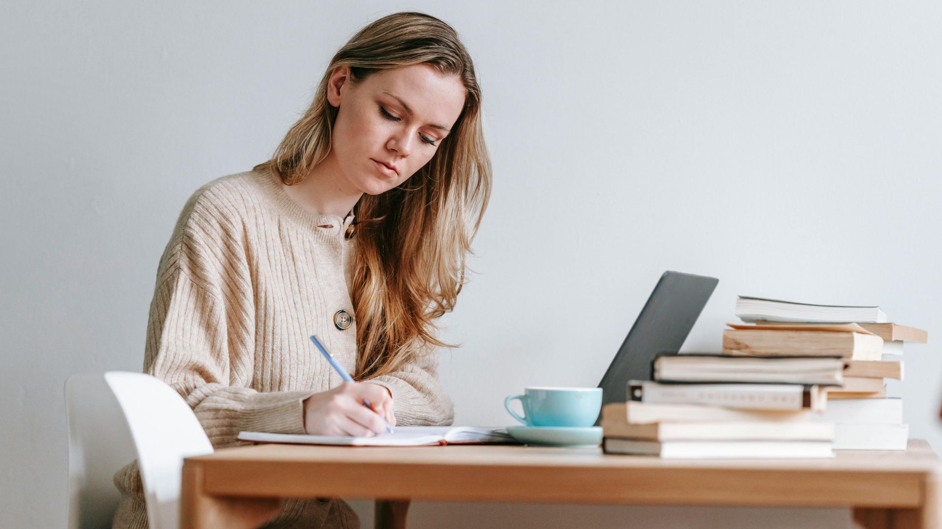 Concentrated female taking notes in planner near heap of many books and cup of strong coffee on white background