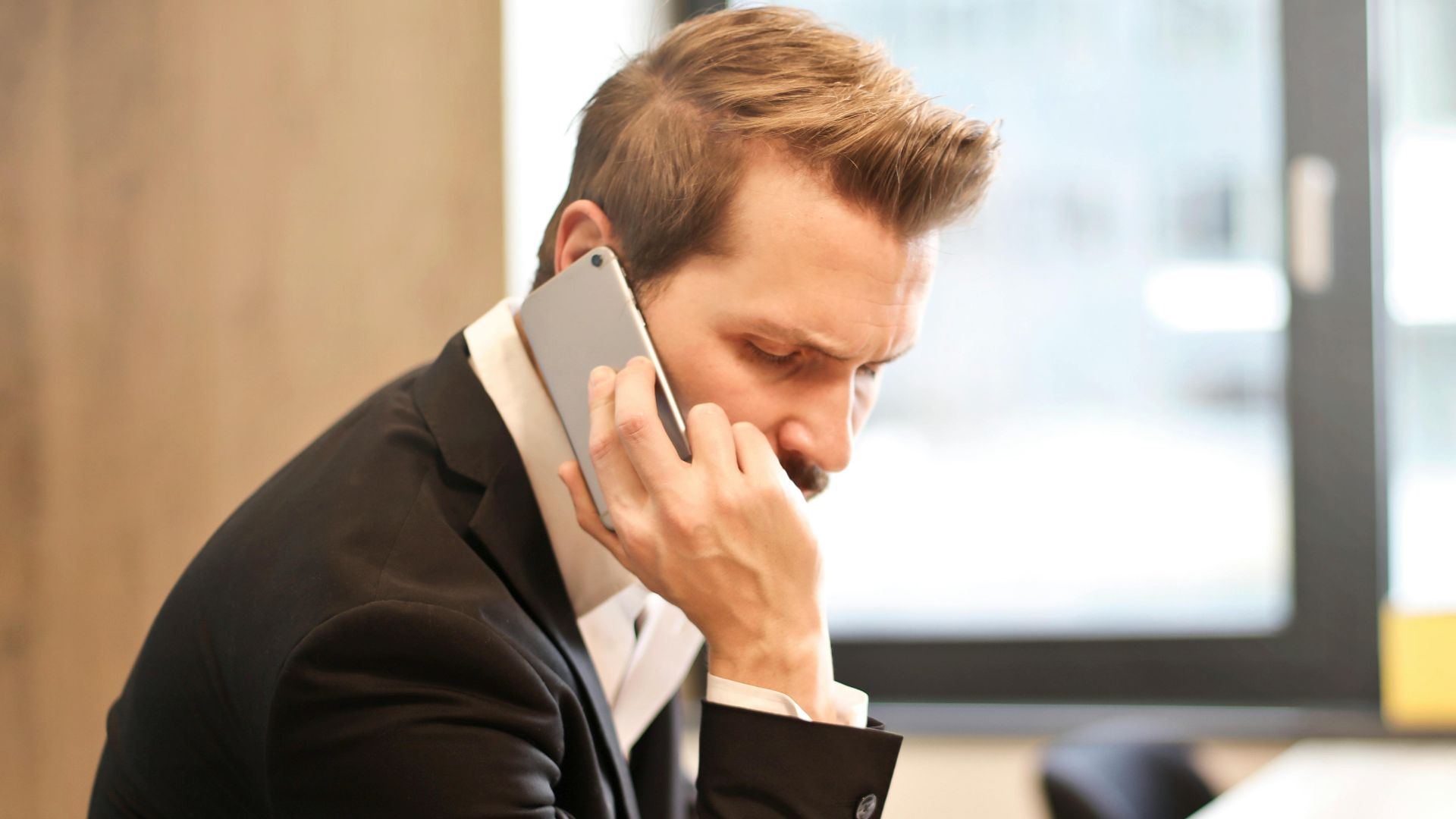 A businessman in a suit making a phone call while working on a laptop in a modern office setting.
