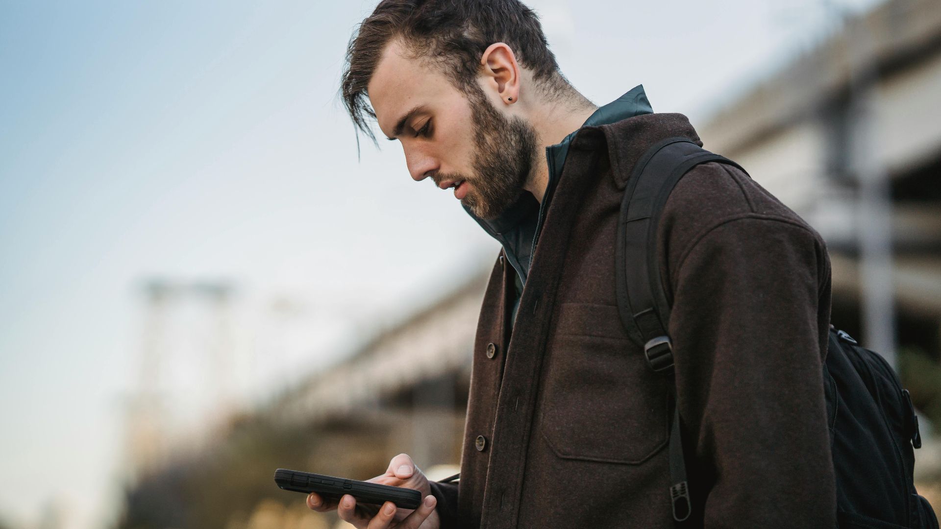 Side view of crop young unshaven male watching cellphone under light sky in town