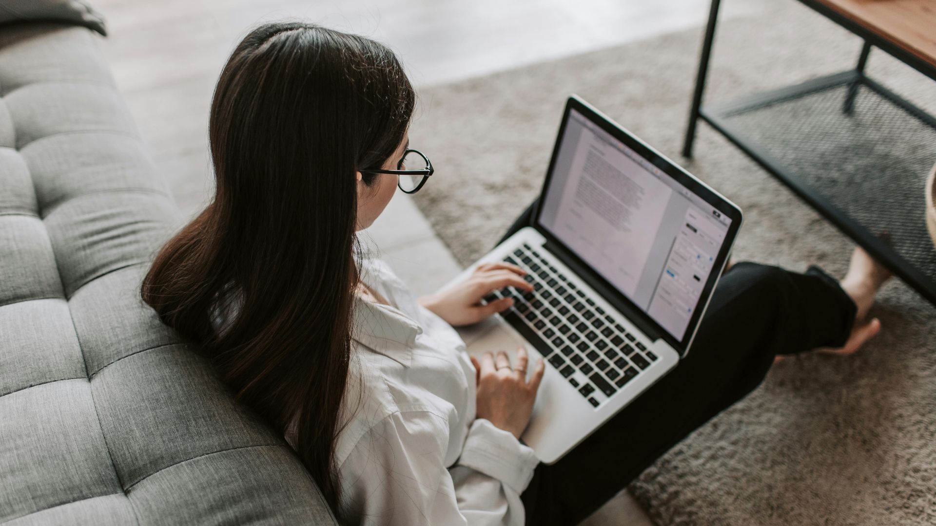 Woman working remotely with a laptop on the floor next to a sofa, enjoying comfortable home office setup.