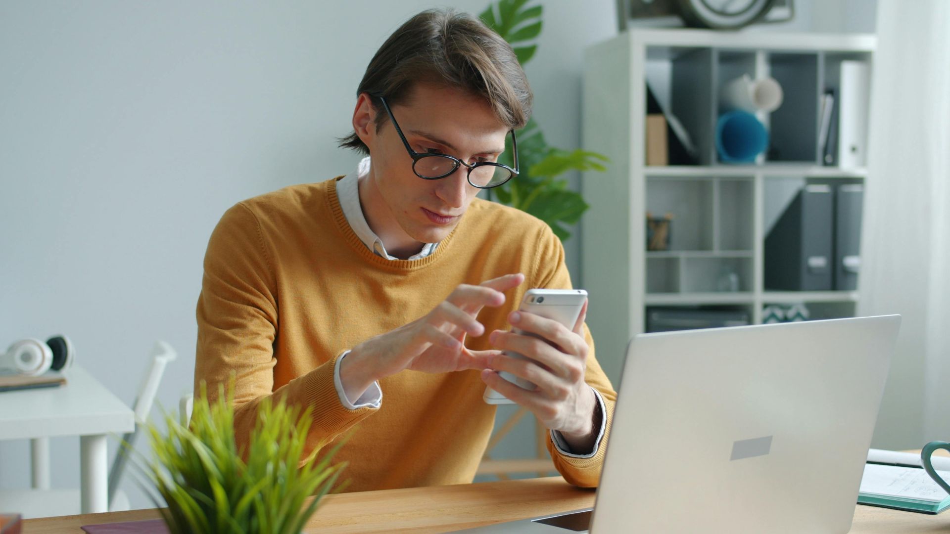 A young adult working on a smartphone and laptop in a modern office.