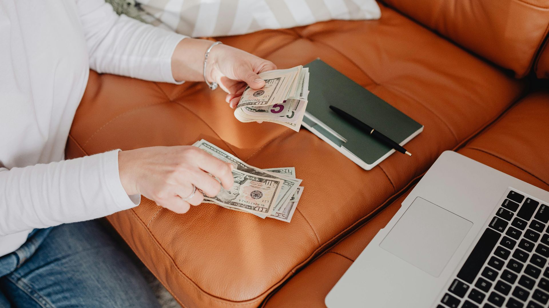 An adult woman sitting on a sofa, counting money with a laptop and notebook nearby, illustrating budget planning.